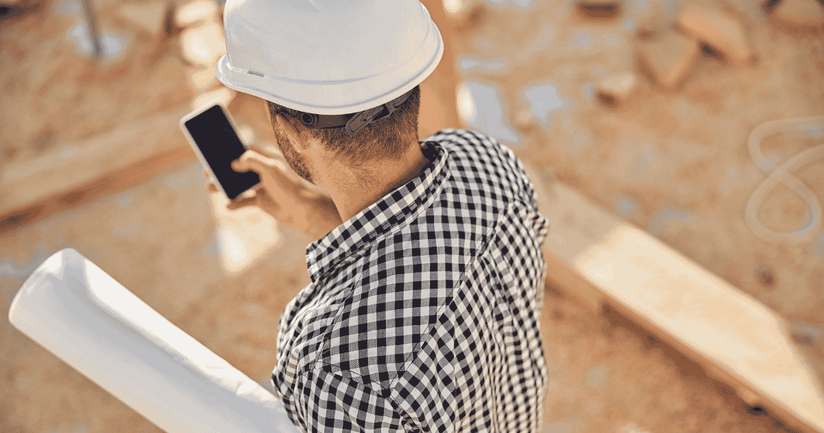 Man looking at smartphone wearing hard hat and holding papers on a construction job site