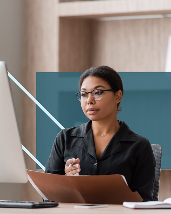 multiracial woman looks at her desktop screen with a folder in her hand.