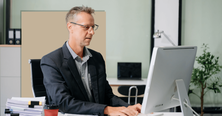 A business man looks at a computer while sitting at a desk with papers and coffee