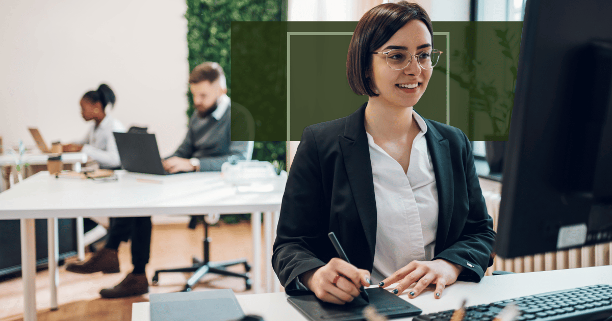 woman looks at her desktop screen while writing notes in office with coworkers in the background. woman looks at her desktop screen while writing notes in office with coworkers in the background.