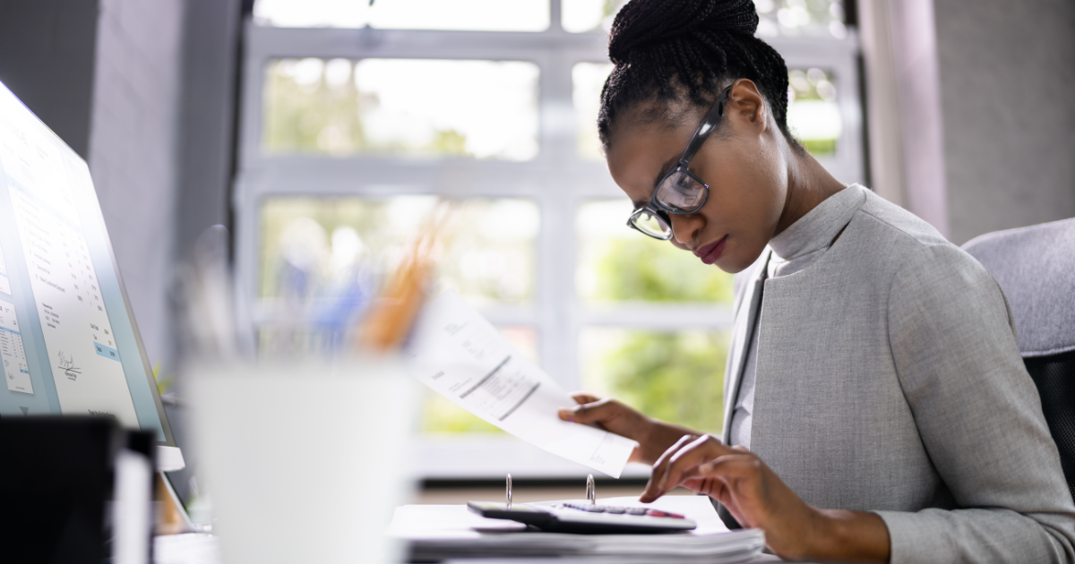 A woman in her office types on her calculator while holding a piece of paper with a desktop in the background A woman in her office types on her calculator while holding a piece of paper with a desktop in the background