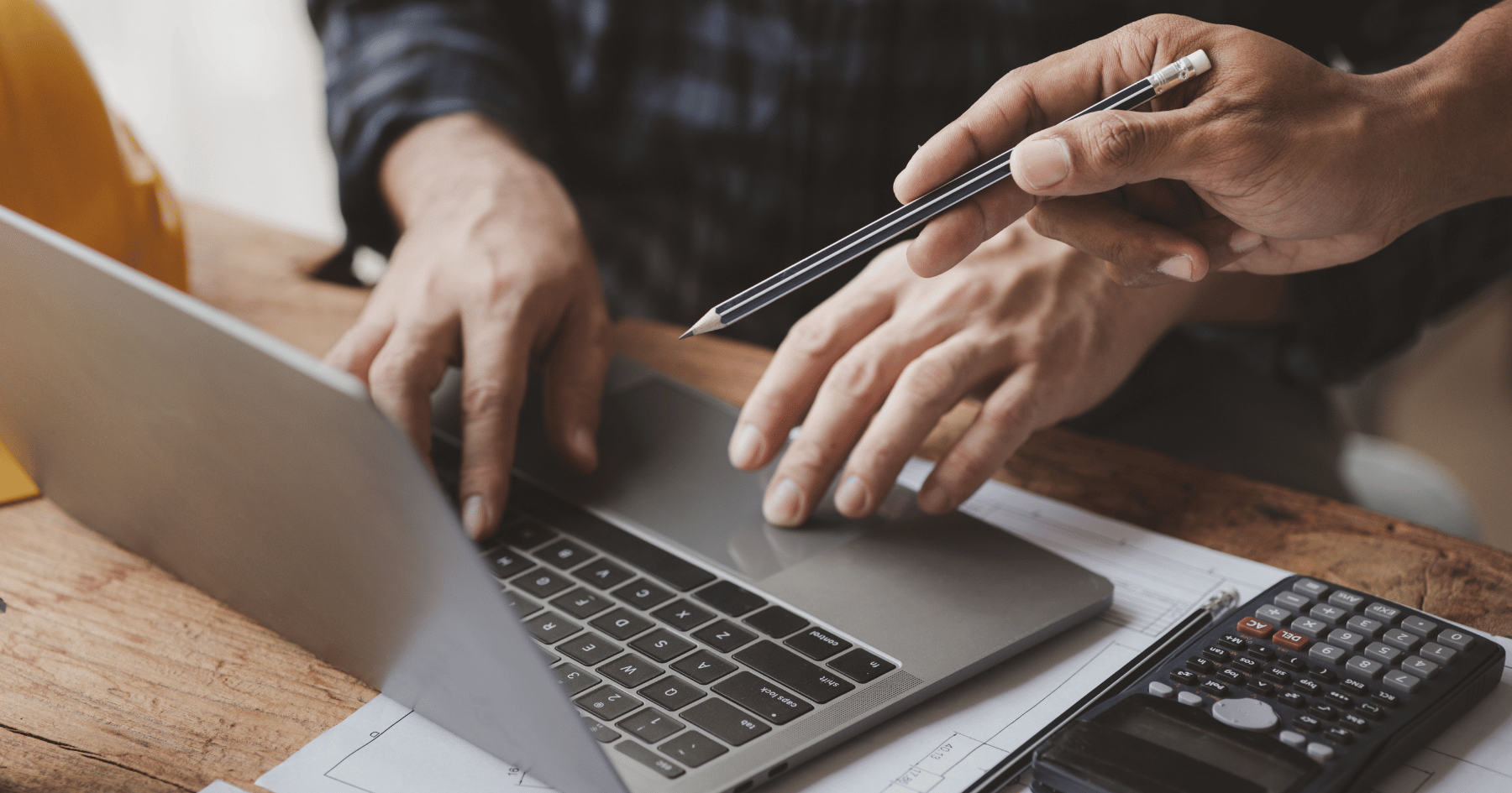 Two people work on a plan in a workplace. A laptop, a calculator, building plans, and a yellow hard hat are on the wooden desk. 