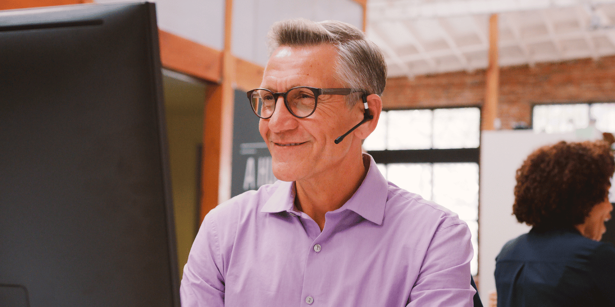 A man talking on a headset smiles at his computer